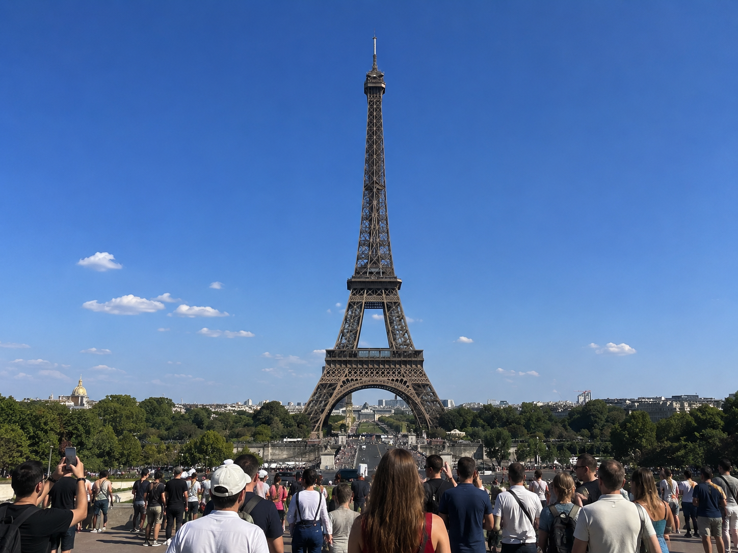 A scenic view of the Eiffel Tower in Paris under a bright blue sky, with a crowd of tourists gathered in the foreground on the Champ de Mars.