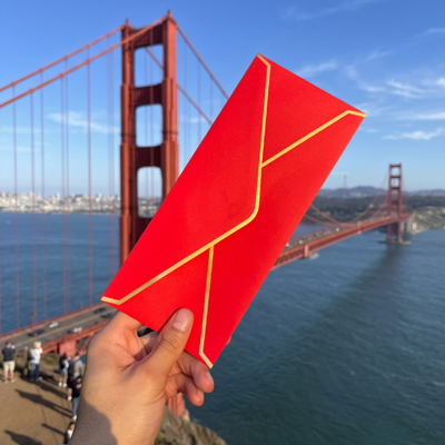 A hand holding a red and gold envelope in front of the Golden Gate Bridge.
