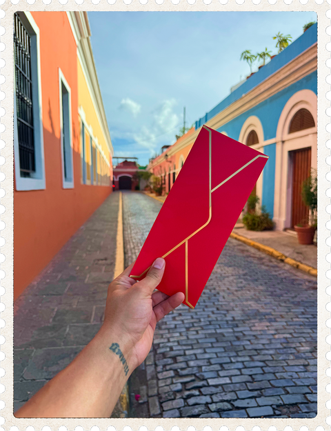 A hand holding a red envelope with gold detailing along a colorful cobblestone street lined with vibrant colonial buildings, framed by a vintage postage stamp border.